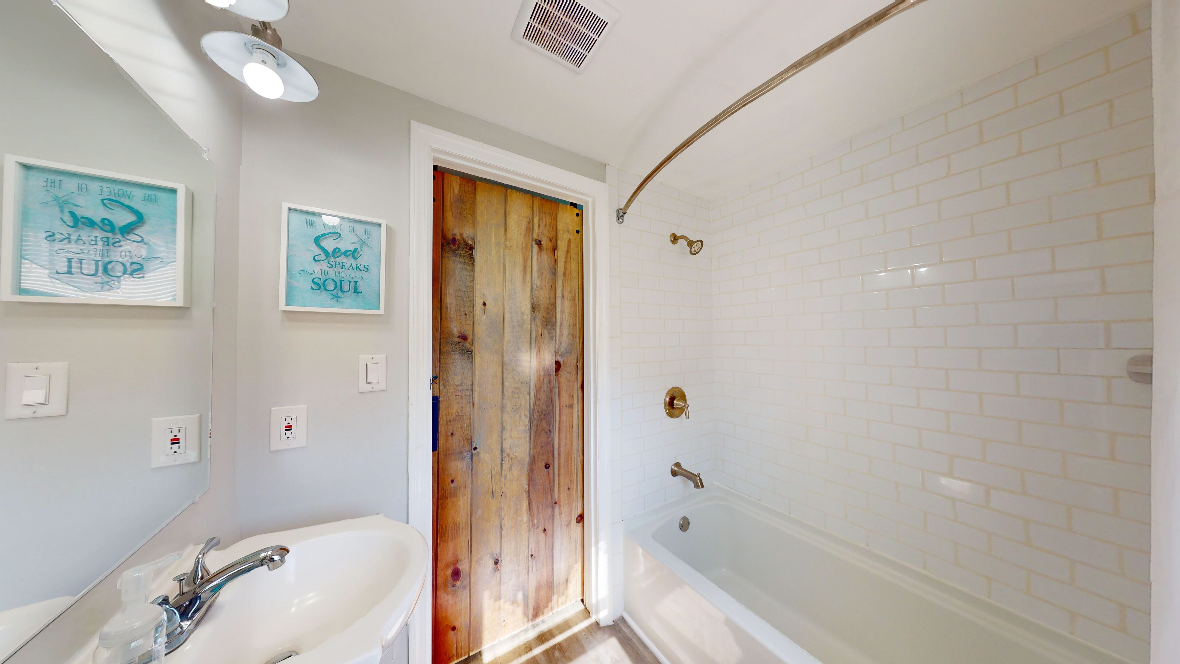 A bathroom with a wooden door, white sink, and a white bathtub. There are two framed quotes on the wall and a light fixture above the sink.