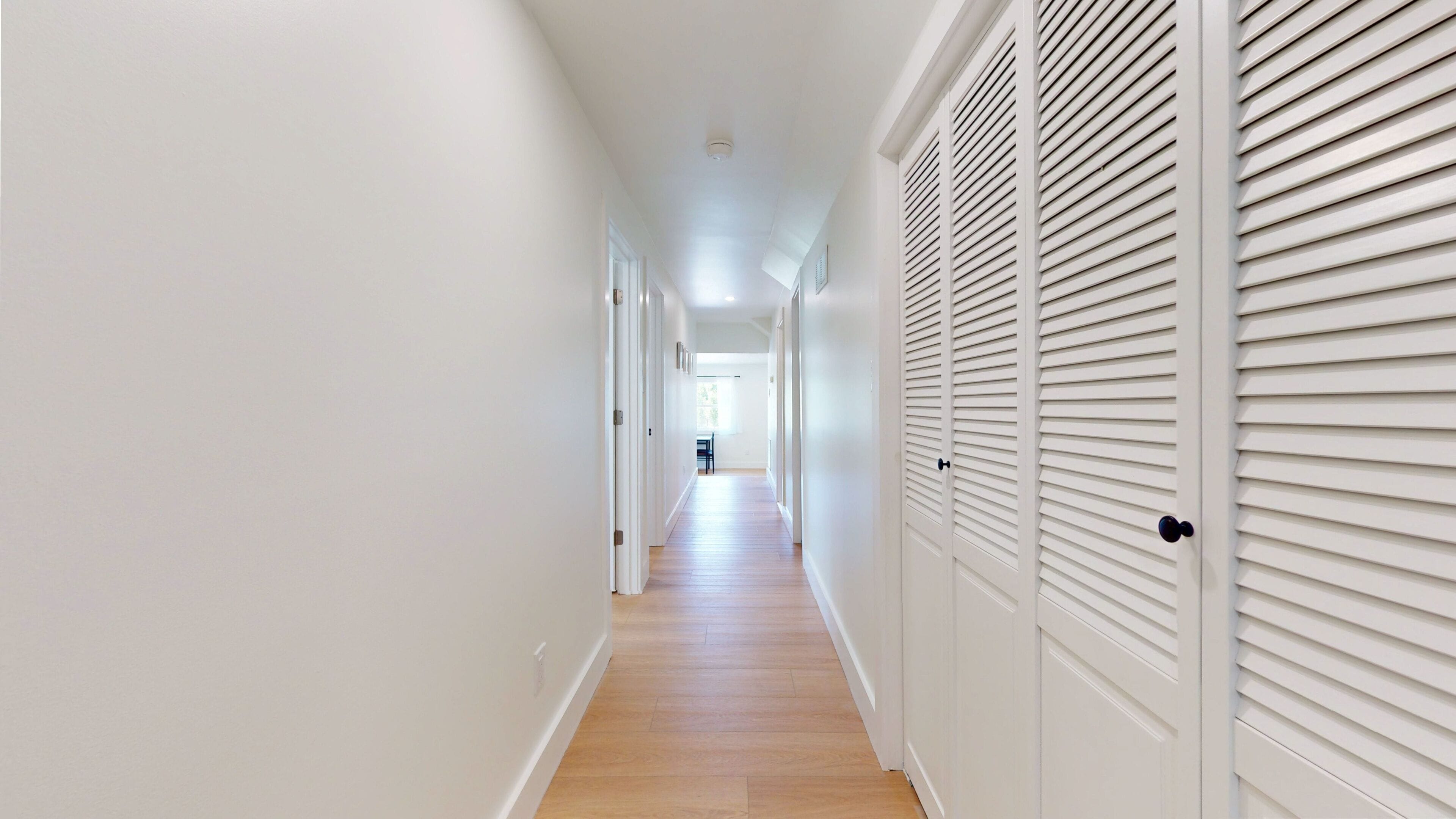 Un couloir aux murs blancs, au plancher en bois et doté d’une porte avec des stores horizontaux.