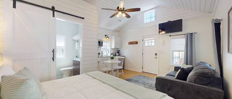 A bedroom with a bed, a ceiling fan, a window, and a view into a kitchen.