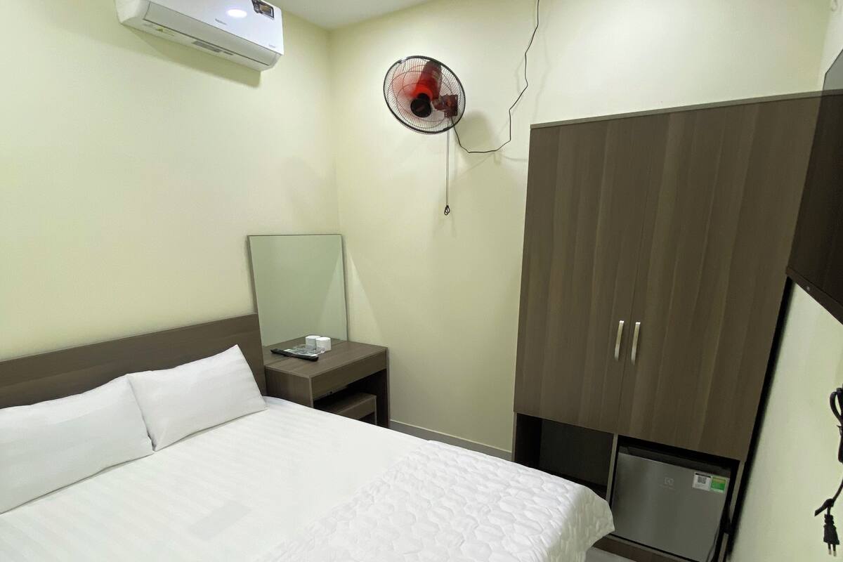 Quarto de hotel com cama, ar condicionado, ventoinha, espelho, guarda-roupa e um pequeno frigorífico.