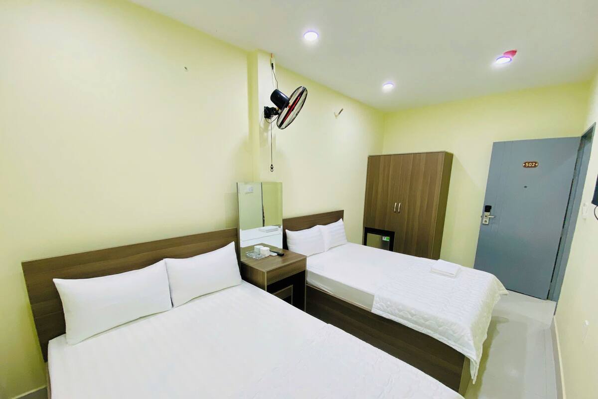 Quarto de hotel com duas camas de solteiro, um guarda-roupa de madeira, um espelho, uma mesa de cabeceira com abajur e um ventilador de teto.