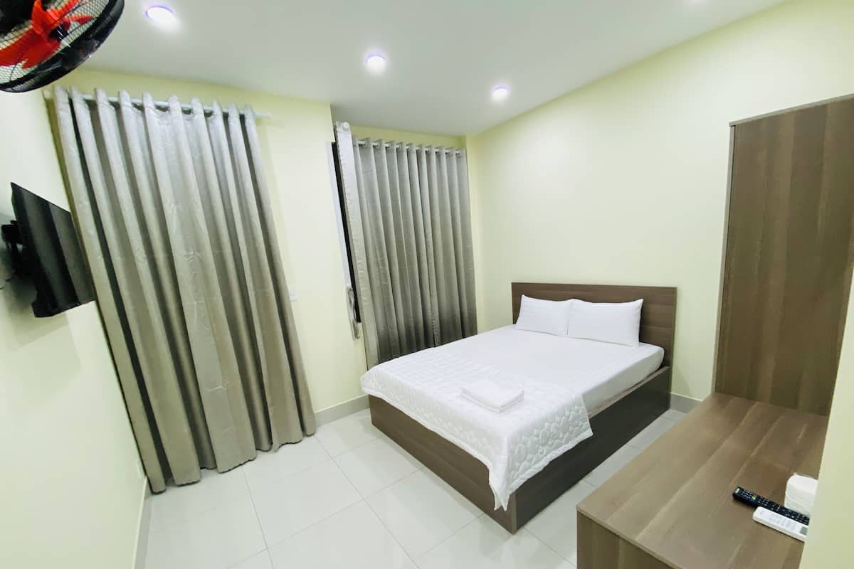 Quarto de hotel com uma cama, cortinas, uma TV fixada na parede e uma secretária de madeira com um comando remoto.