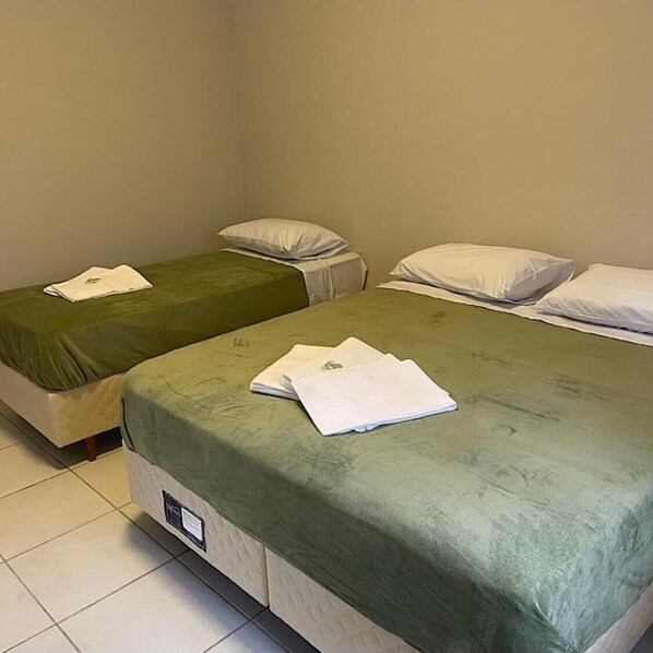 Two single beds with green bedspreads and white pillows, a bedside table with a white cloth, and a tiled floor.