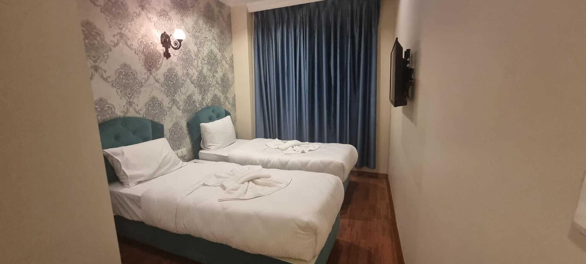 Une chambre d’hôtel avec deux lits, un papier peint à motifs, un rideau bleu et un téléviseur fixé au mur.