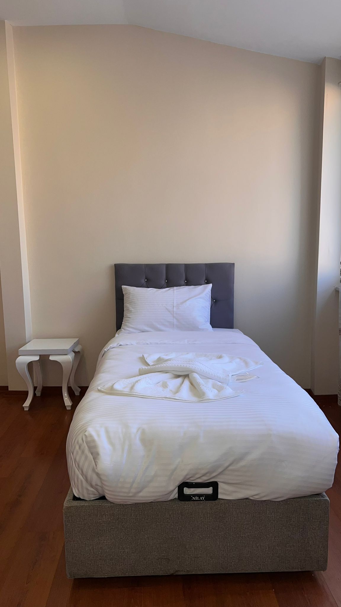 Un lit bien fait, avec une tête de lit grise et un oreiller blanc, une petite table de chevet blanche et un matelas gris posé sur le sol.