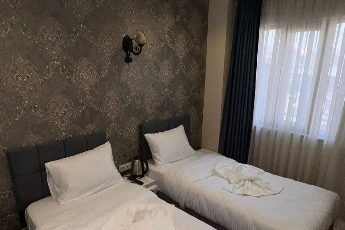 A hotel room with two beds, a patterned wallpaper, a wall-mounted lamp, and a window with curtains.