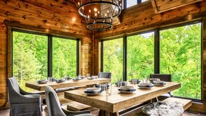 A wooden cabin with a dining area featuring a large table set for four, surrounded by large windows offering a view of lush greenery. - Heated Pool/HotTub/Games/Fire/Sleep 20+ (Sevierville)