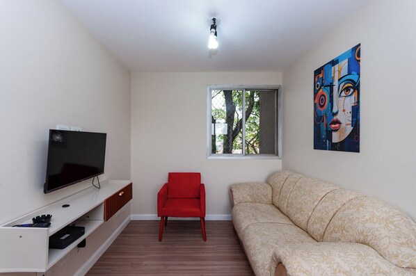 A living room with a flat-screen TV, a red chair, a beige sofa, a window with a view of trees, and a colorful painting on the wall. - Ad disabled Ad disabled   (Lapa de Baixo São Paulo)