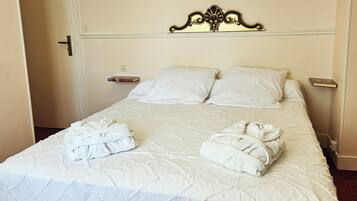 A neatly made bed with white bedding and two towels, a wall-mounted light fixture, and a door to the left.