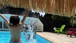 Ein Schwimmbecken mit einer Person, die Wasser spritzt, umgeben von einer Holzterrasse und zwei grünen Stühlen.