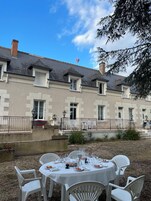 Une cour avec une table dressée pour le repas, entourée de chaises blanches.
