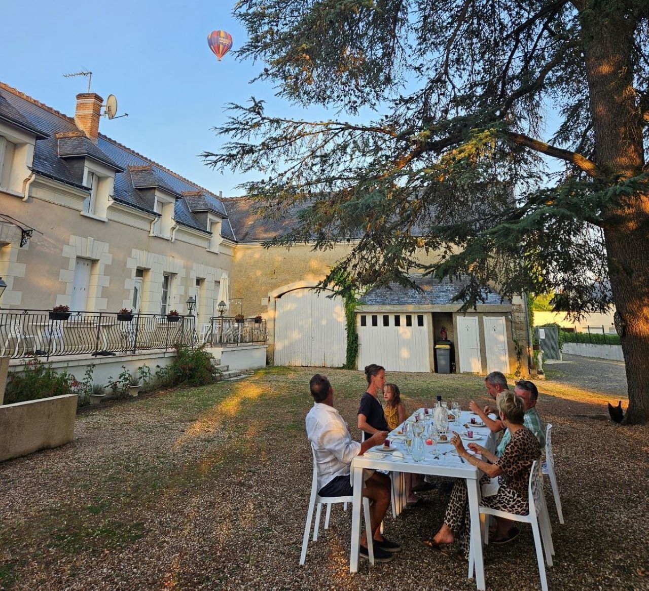 Un grupo de personas cenando al aire libre en una mesa con mantel blanco, rodeado de un jardín y un edificio con tejado de baldosas.