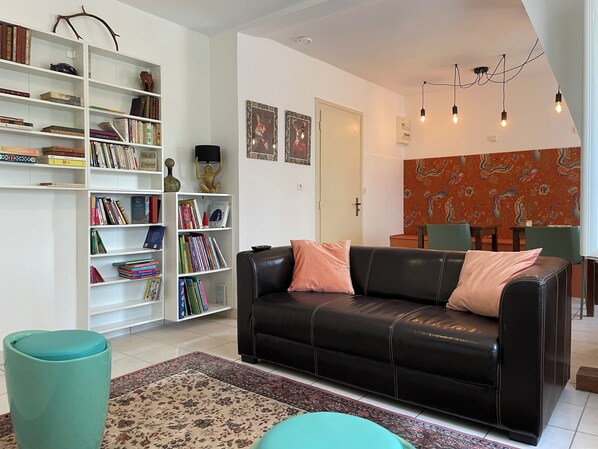 A living room with a black leather sofa, a bookshelf, a patterned rug, and a decorative wall hanging. - Aux Chambres des Dames (Chisseaux)