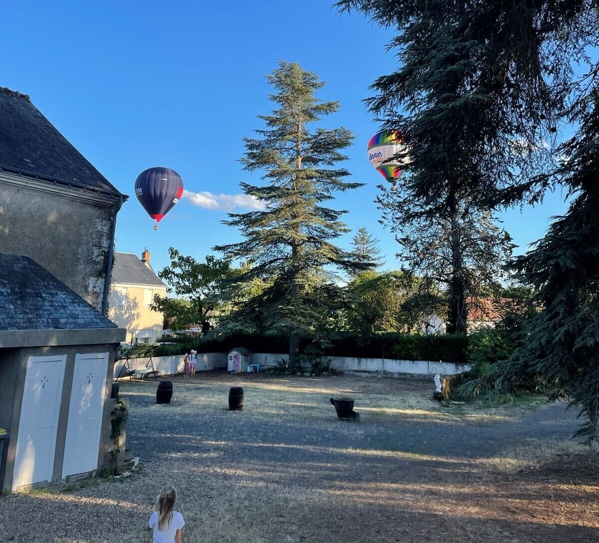 A courtyard with a large coniferous tree, a building with a sloped roof, and two hot air balloons in the background.