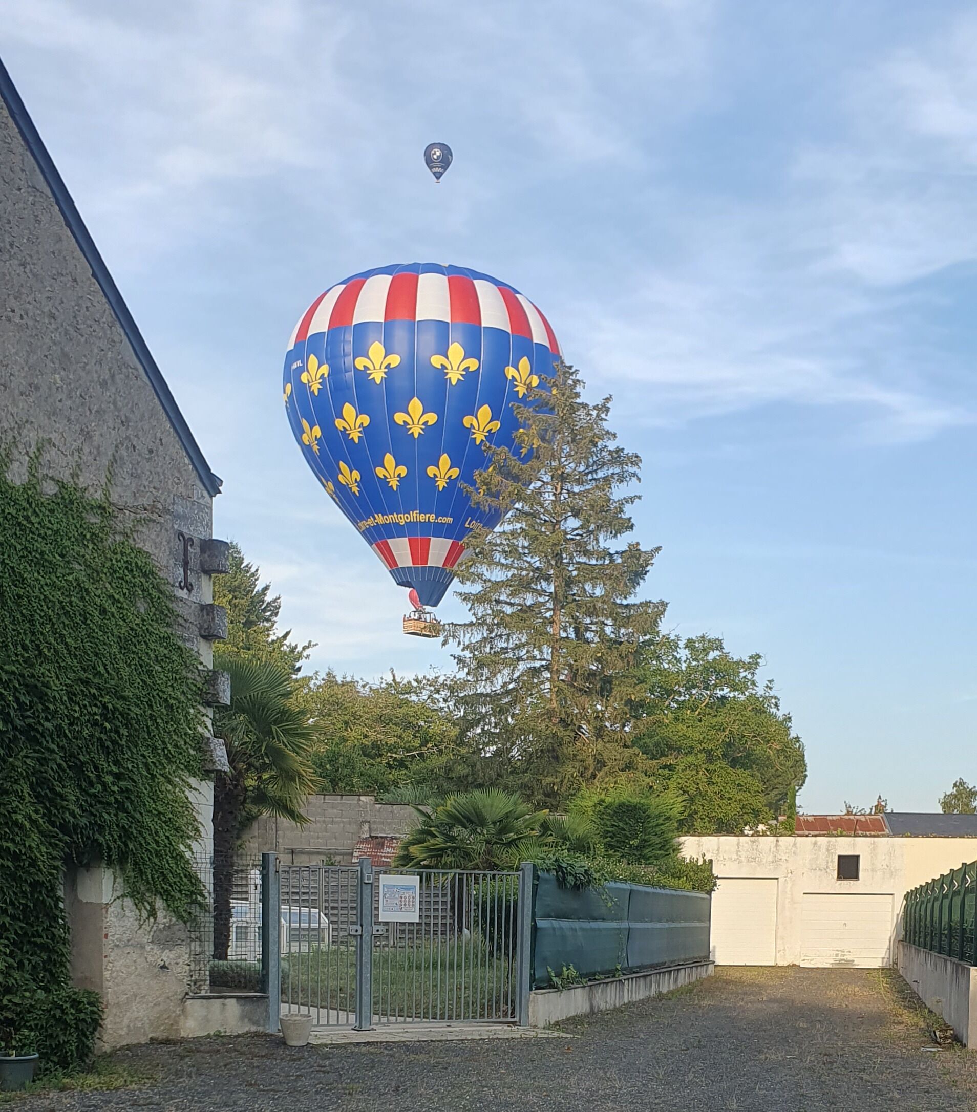 A hot air balloon with a blue, red, and white striped pattern and yellow fleur-de-lis symbols, flying above a building with ivy-covered walls and a metal gate.