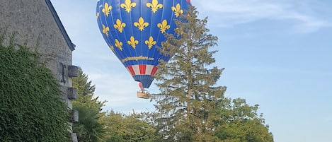 Un globo aerostático con un diseño a rayas azul, rojo y blanco y símbolos de flores de lis amarillas, volando sobre un edificio con muros cubiertos de hiedra y una reja de metal.