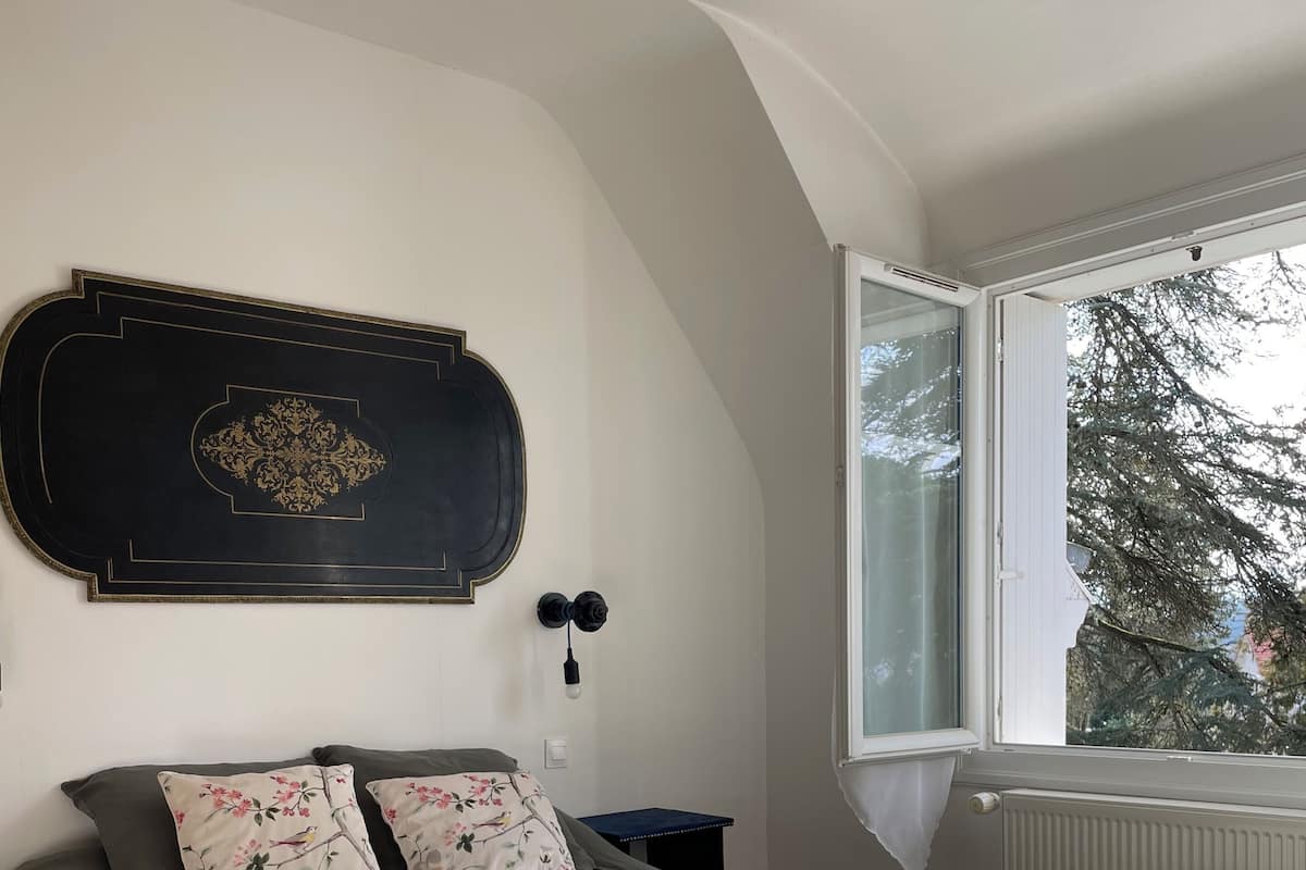 Une chambre à coucher comprenant un lit, une table de chevet, une lampe fixée au mur et une fenêtre donnant sur des arbres.