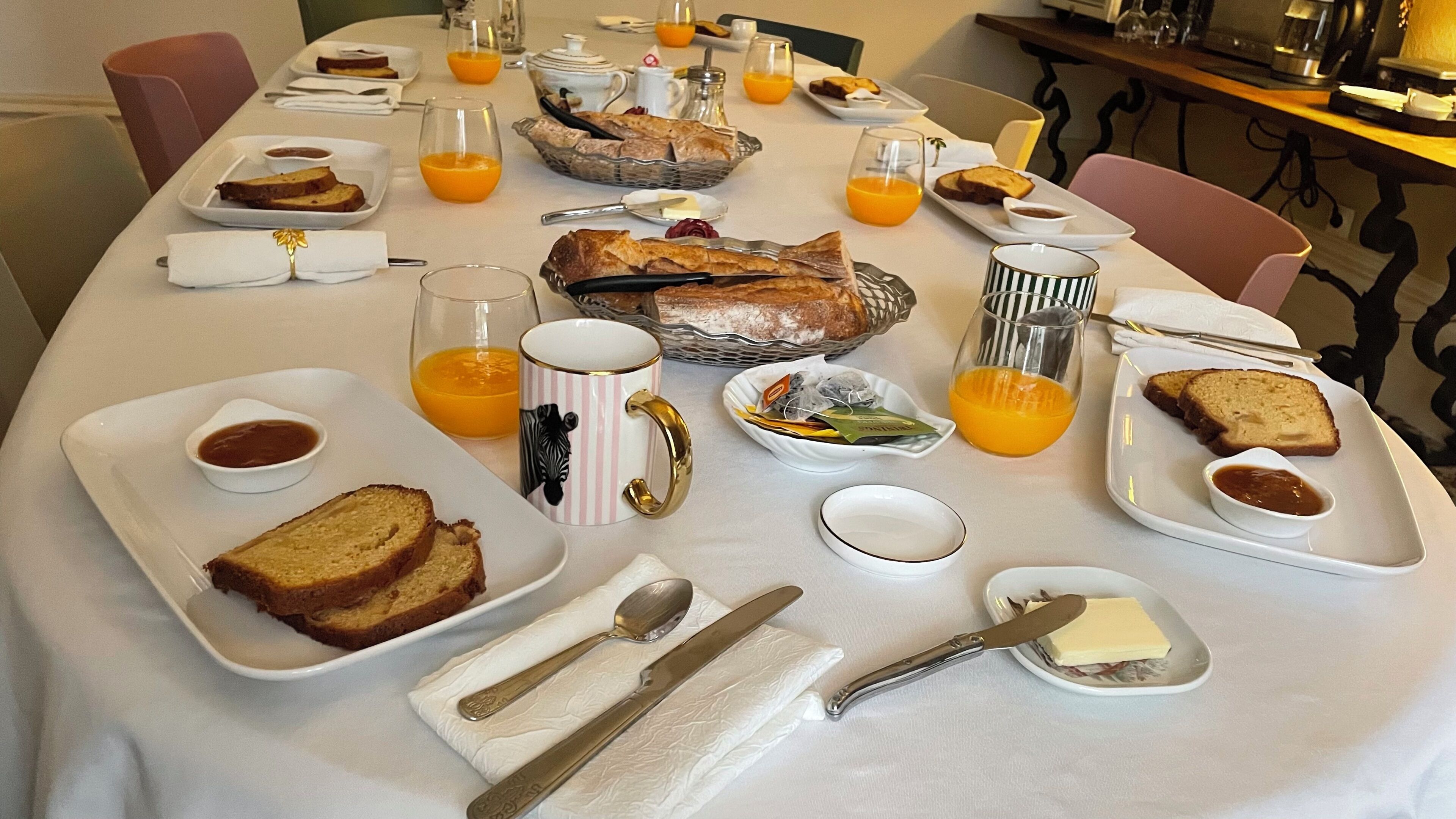 Une table de petit-déjeuner dressée avec du pain, du beurre, de la confiture, du thé et du jus d’orange.