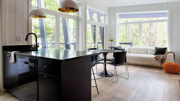 A modern kitchen with a central island, black chairs, a dining table, and a sofa with a view of the outdoors. - Fauna Bluff, A cliffside 2-bedroom modern cottage overlooking a bubbling stream. (Loudonville)