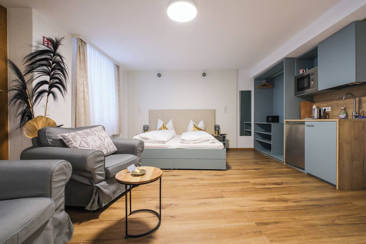 Une chambre d’hôtel moderne avec un canapé, une petite table ronde, un lit avec une literie blanche, un plancher en bois, un coin cuisine équipé d’un four à micro-ondes et une plante suspendue.