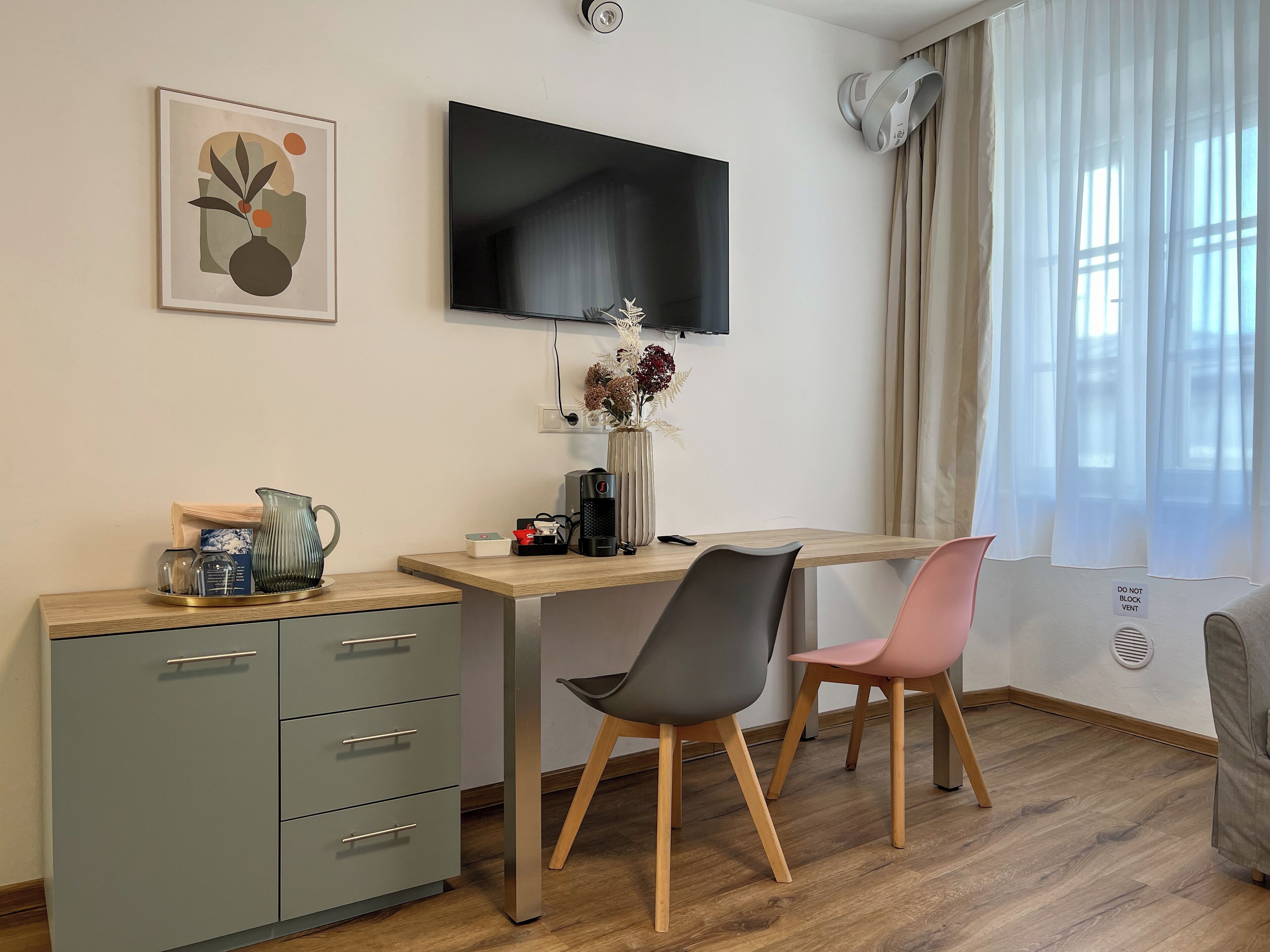 A modern room with a desk, two chairs, a TV, a coffee maker, a vase with flowers, and a cabinet.