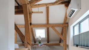 A room with exposed wooden beams, a desk with chairs, a window with blinds, and an air conditioning unit. - Apartment Salzburg - Bergstrasse (Salzburg)