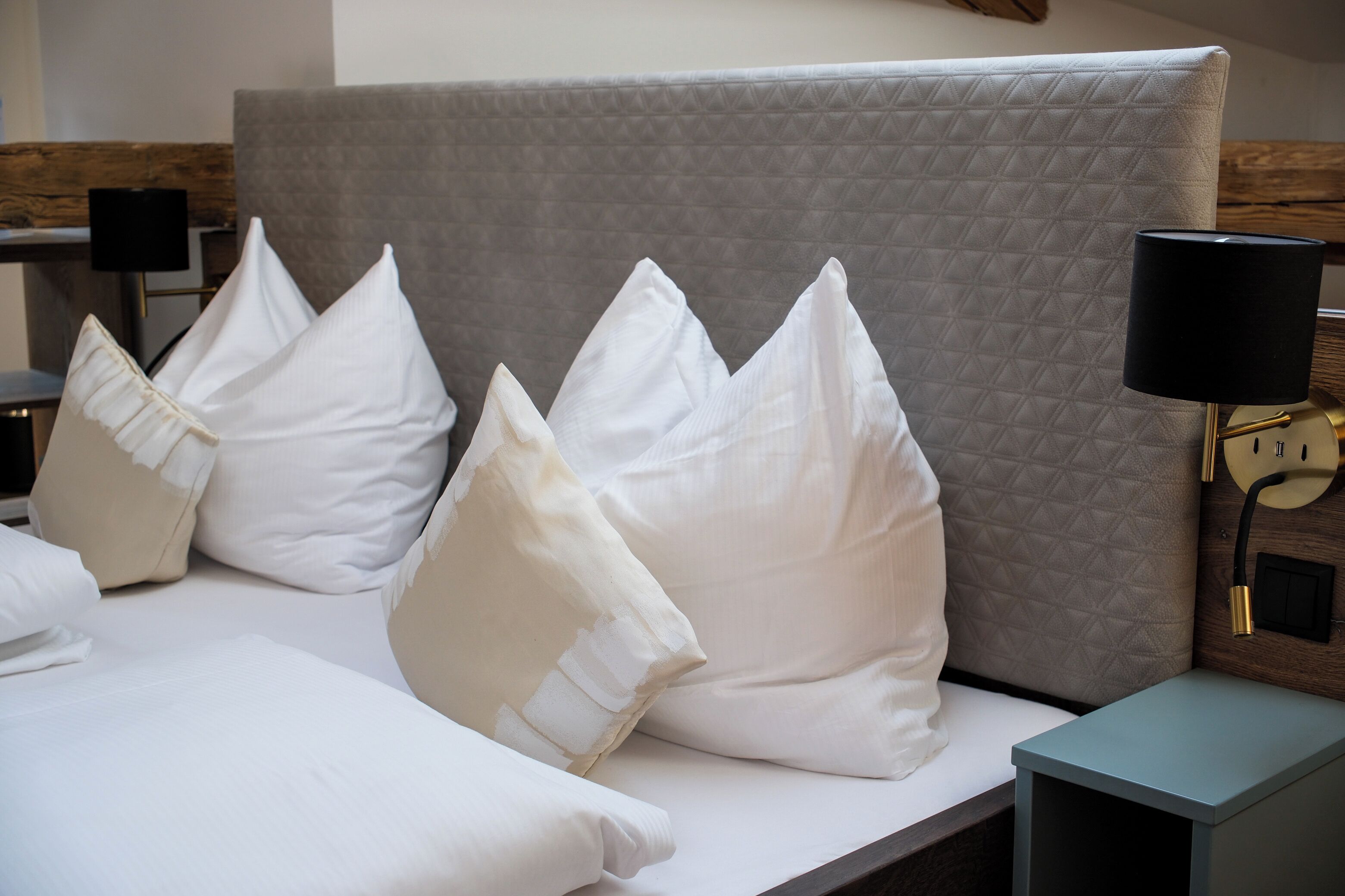 A neatly made bed with white linens and pillows, a grey headboard, and a bedside table with a lamp.