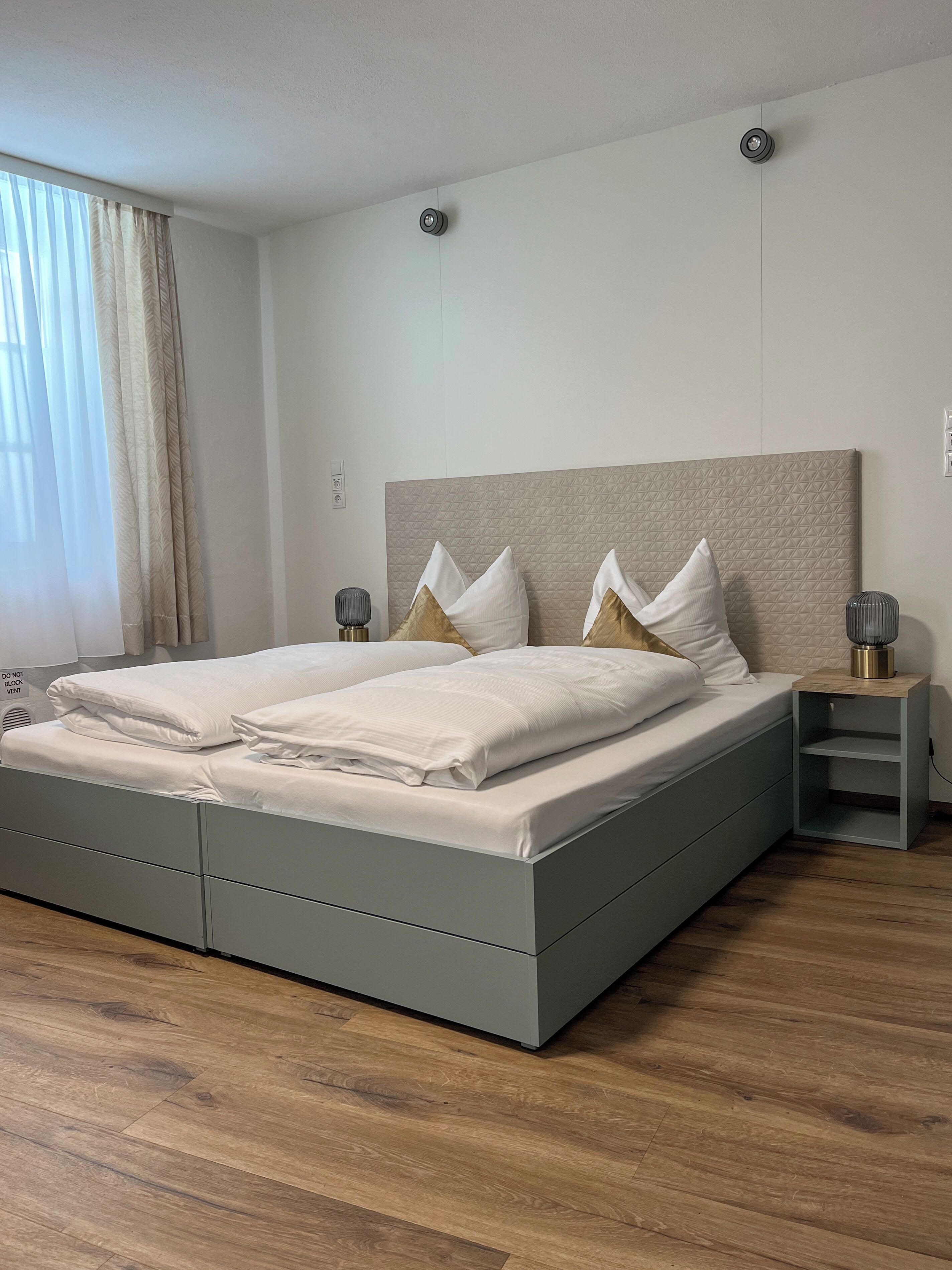 A modern bedroom with a grey bed, white linens, a wooden headboard, and a bedside table with a lamp.