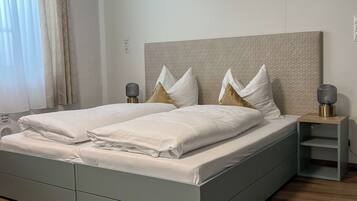 A modern bedroom with a grey bed, white linens, a wooden headboard, and a bedside table with a lamp.