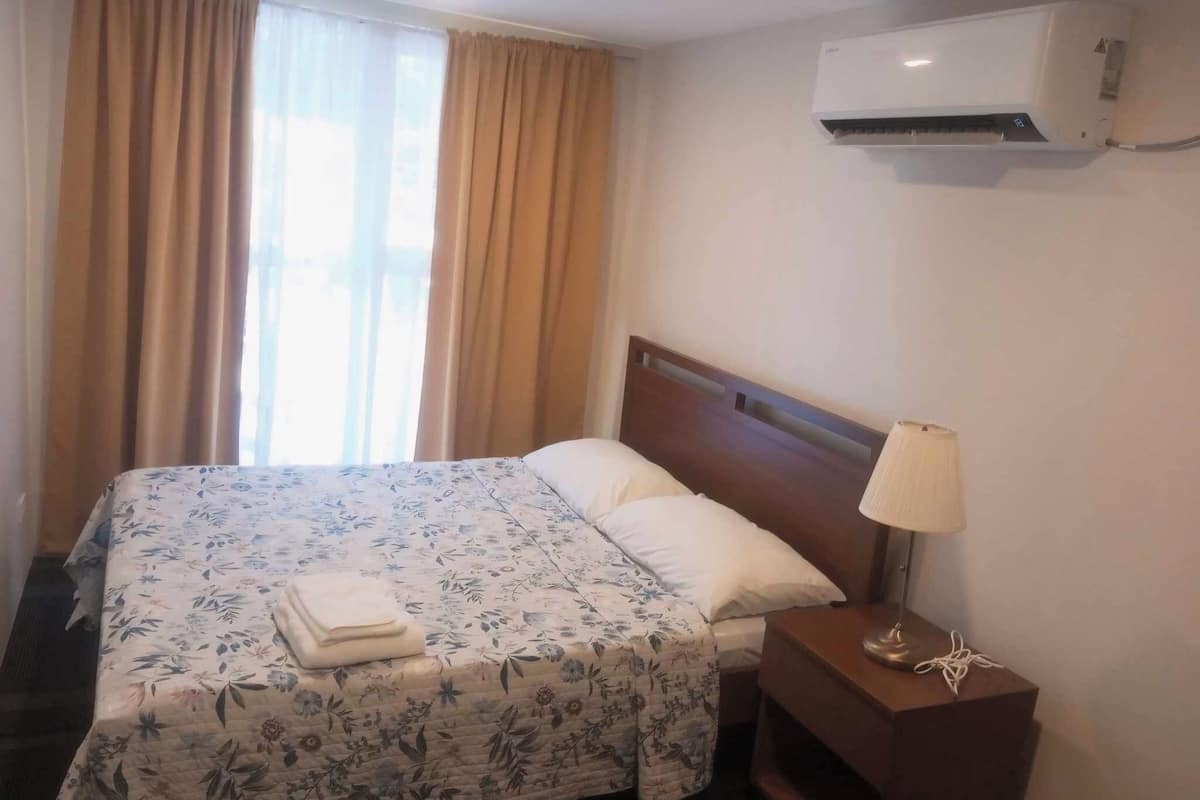 A hotel room with a bed, a nightstand, a lamp, and an air conditioning unit.
