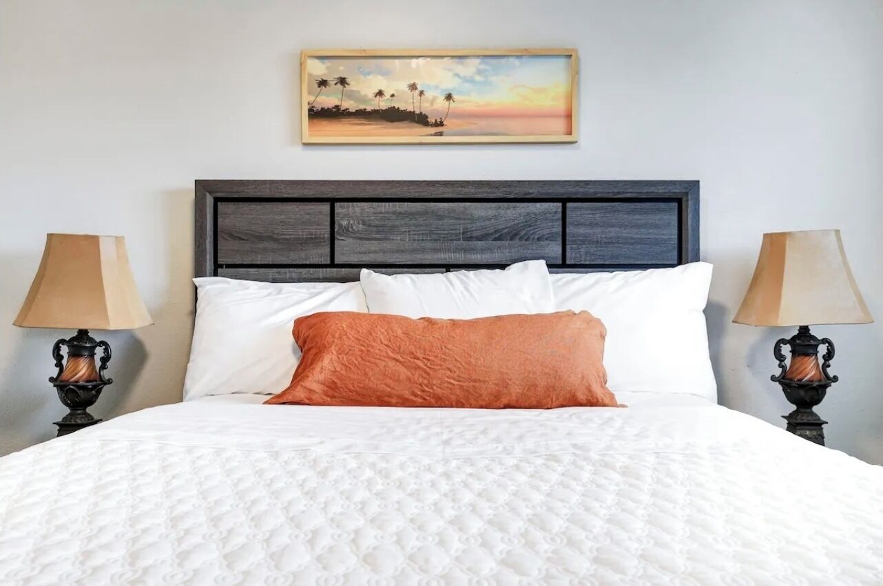 Una cama bien hecha con ropa de cama blanca y naranja, un cabecero oscuro y un cuadro enmarcado encima.