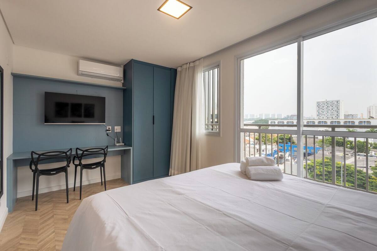 Une chambre d’hôtel équipée d’un lit, d’un bureau, de deux chaises, d’une télévision et d’une grande fenêtre donnant sur la ville.