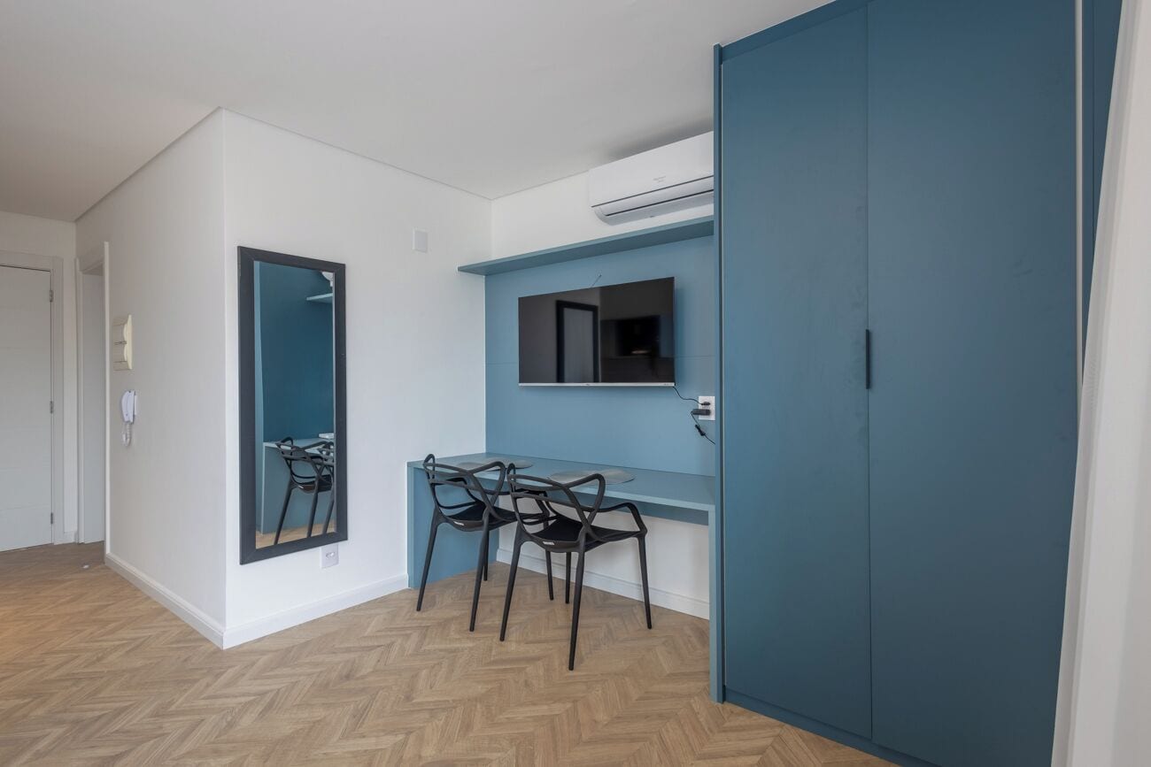 Quarto com piso de madeira em padrão herringbone, uma escrivaninha com duas cadeiras, um espelho grande e uma parede azul.