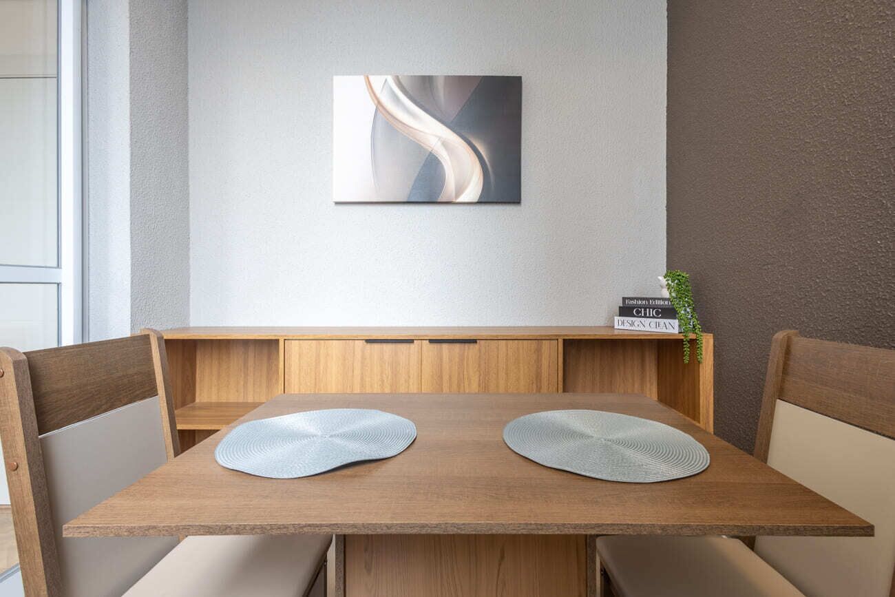 A modern dining area with a wooden table, two cushioned seats, a wall-mounted abstract painting, and a small plant on a shelf.