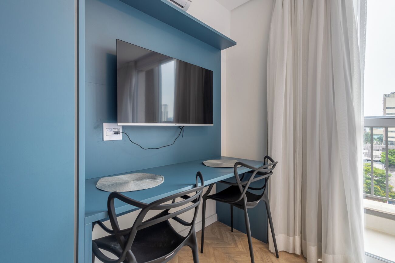A modern room with a blue accent wall, a flat-screen TV mounted above a small table and two chairs, and a large window with white curtains.