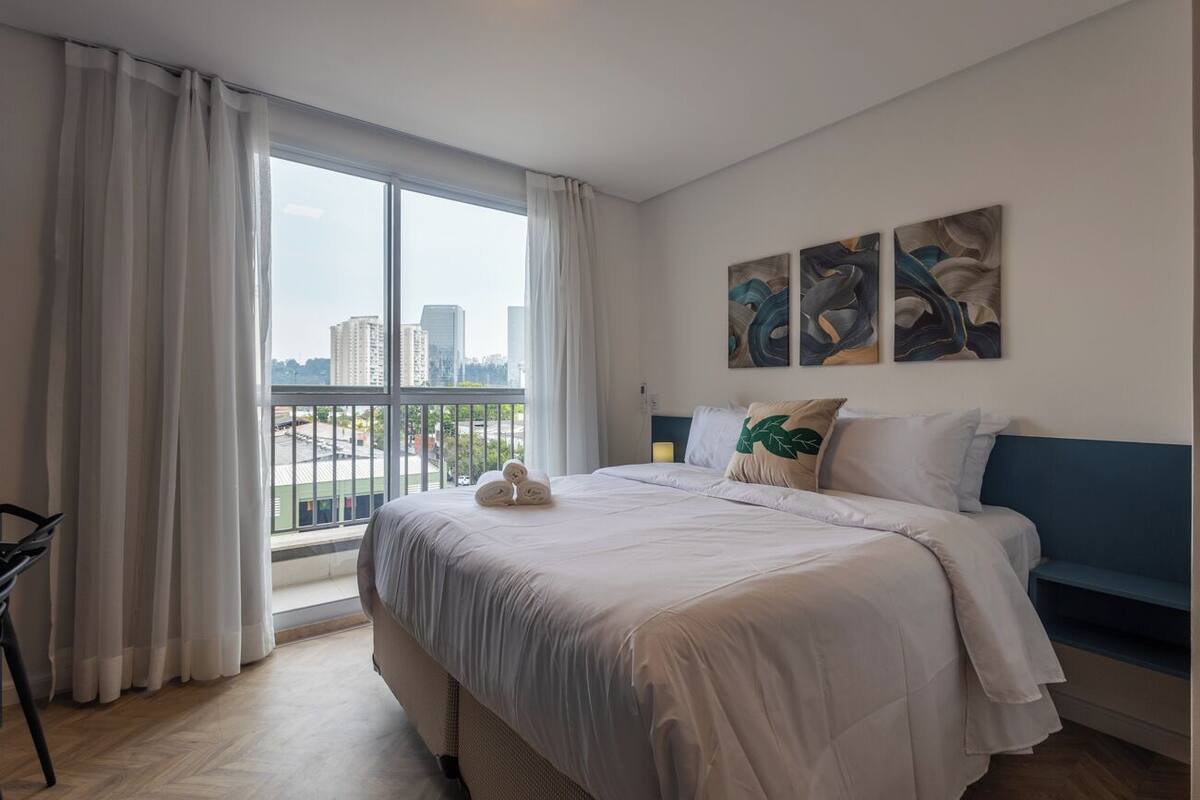 Une chambre d’hôtel moderne dotée d’un grand lit, offrant une vue sur la ville grâce à de larges fenêtres du sol au plafond, et agrémentée d’œuvres d’art murales abstraites.