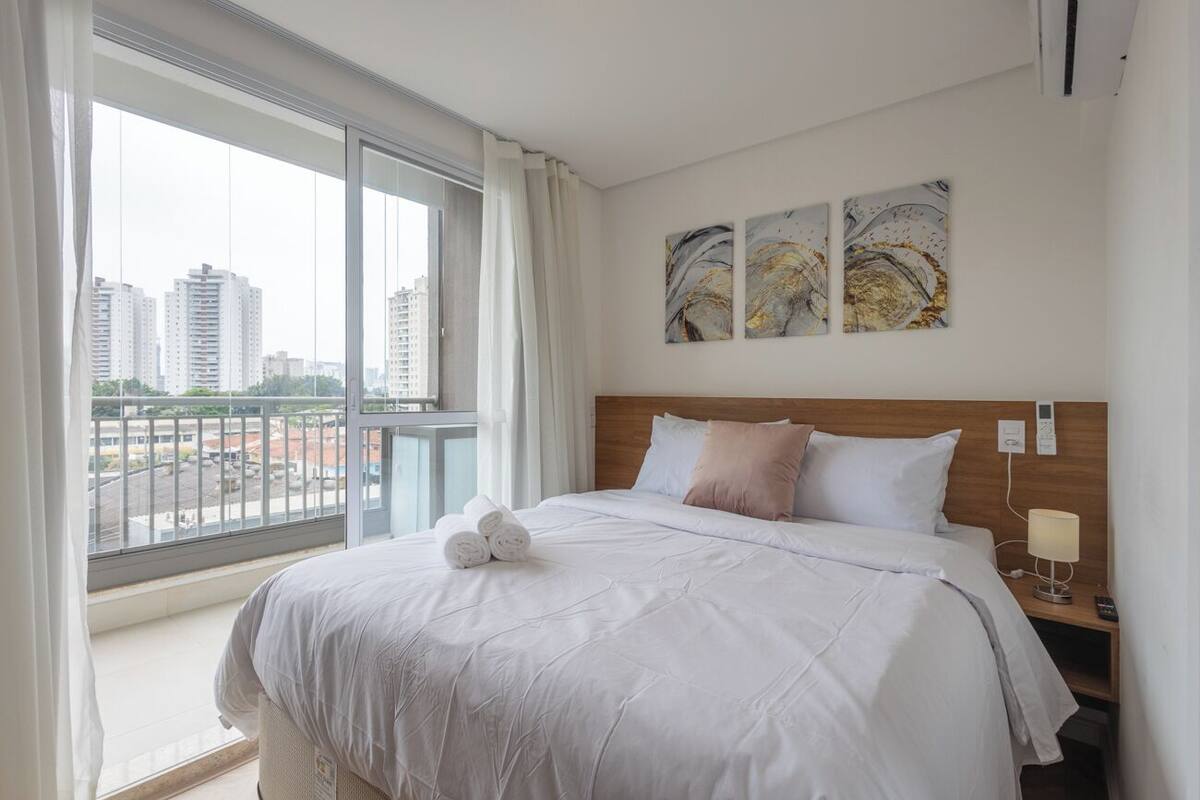 Une chambre d’hôtel avec une grande fenêtre, un lit recouvert de linge de lit blanc et deux tableaux abstraits accrochés au mur.