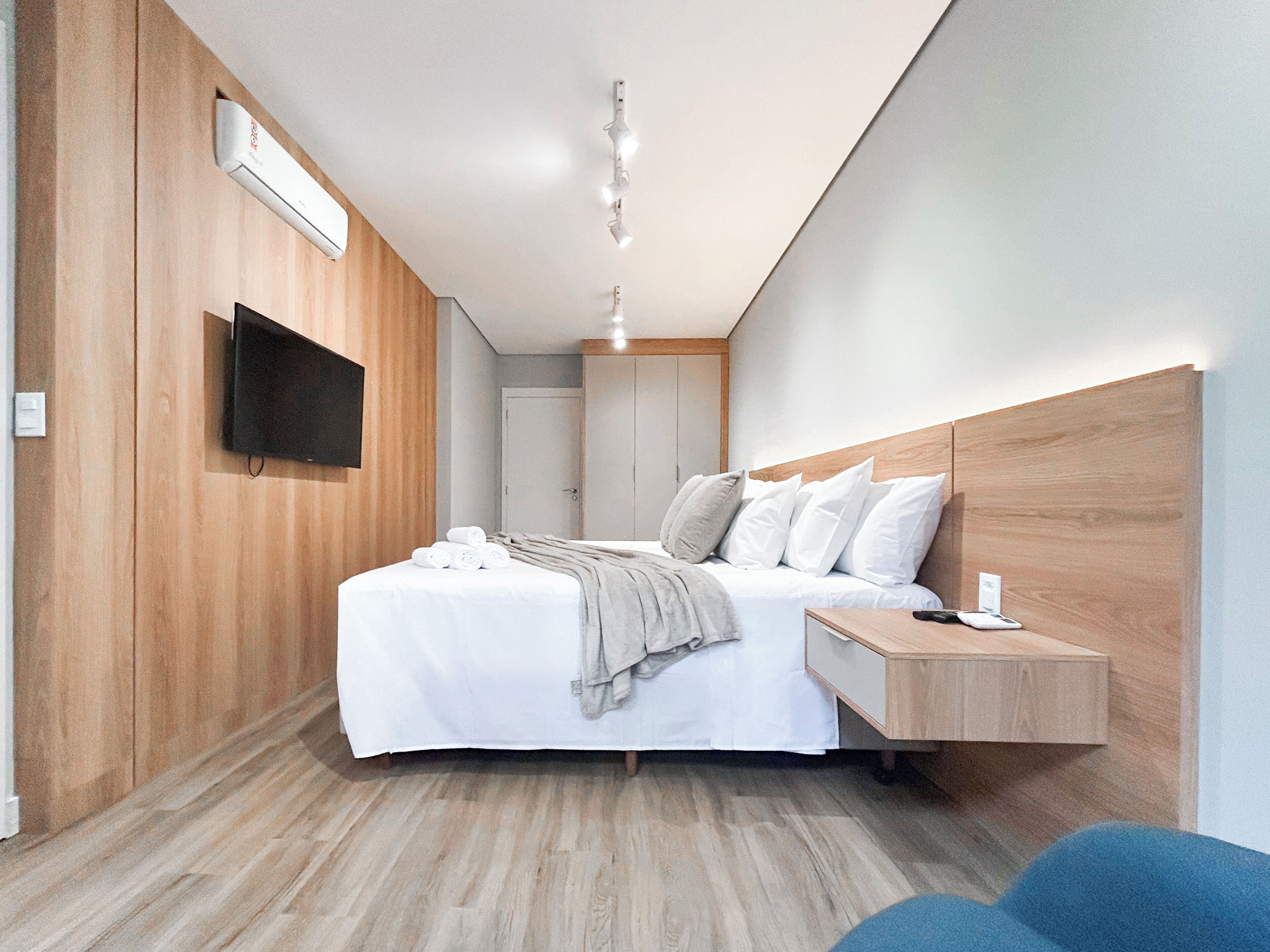 A modern hotel room with a wooden headboard, a flat-screen TV mounted on the wall, a bedside table with a lamp, and a neatly made bed.