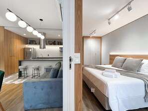 A modern hotel room with a bed, a kitchenette, a seating area with stools, and a wooden accent wall.