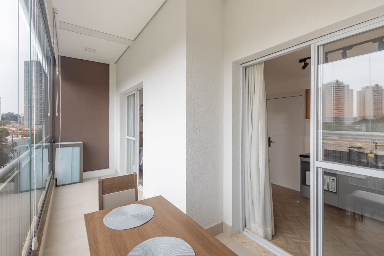 Un balcon avec une table et des chaises pour prendre les repas, une porte-fenêtre coulissante donnant sur une cuisine, et une vue sur de hauts immeubles.