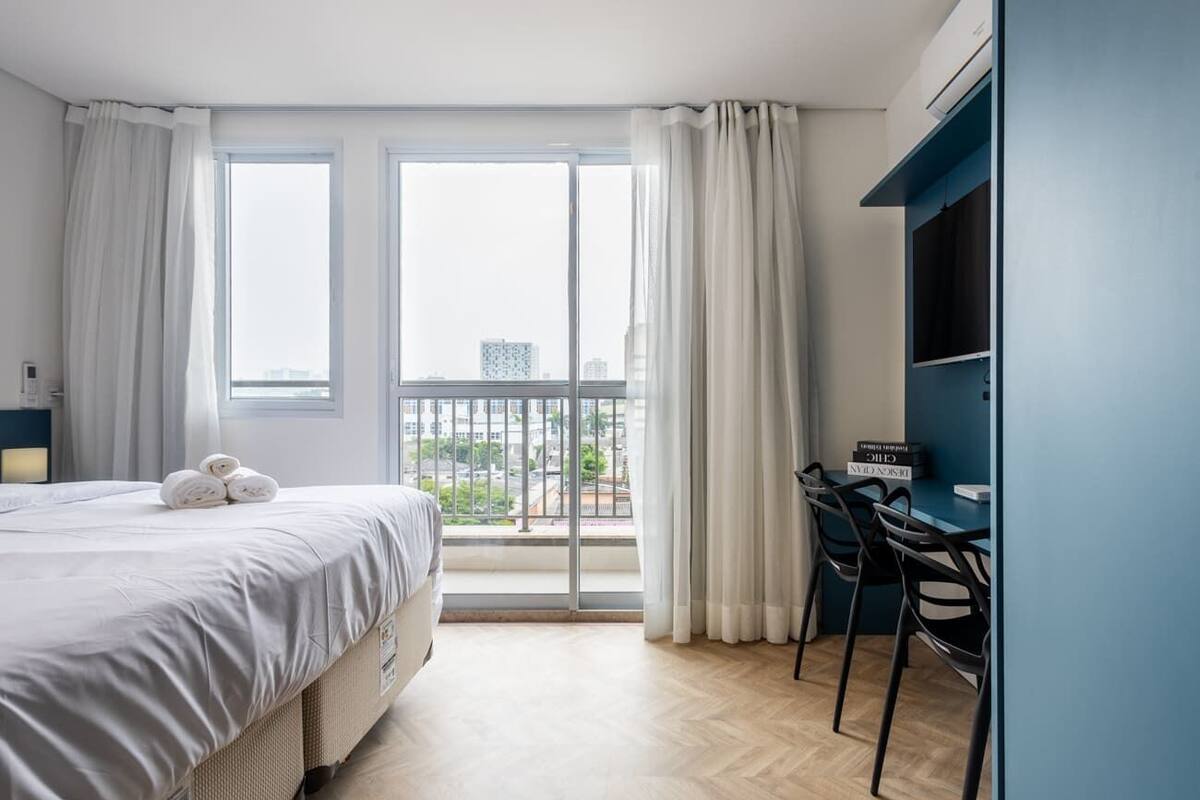 Une chambre d’hôtel moderne dotée d’un grand lit, d’un bureau avec des chaises et offrant une vue sur la ville grâce à de grandes fenêtres.