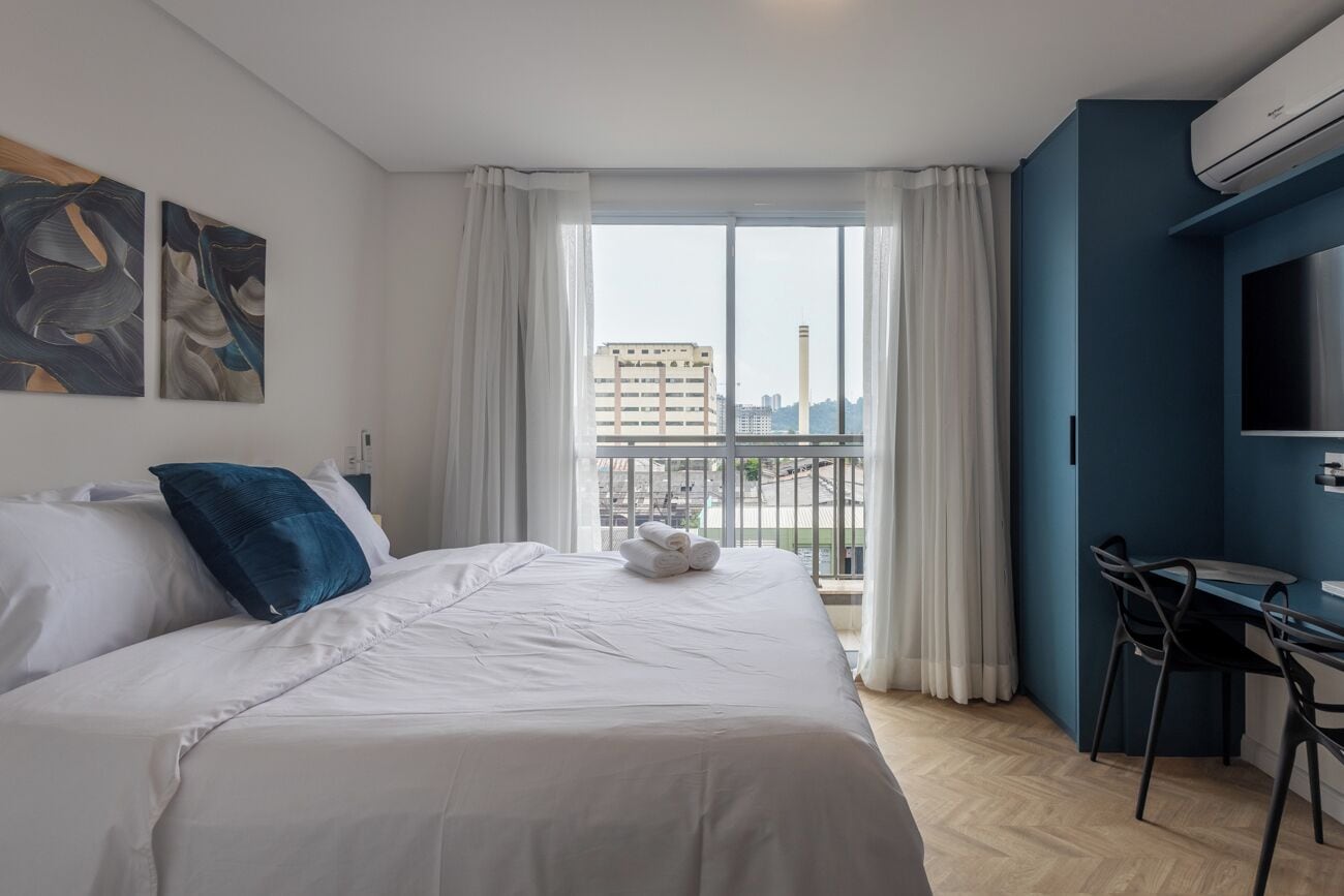 A modern hotel room with a large bed, a desk, and a view of the city through large windows.
