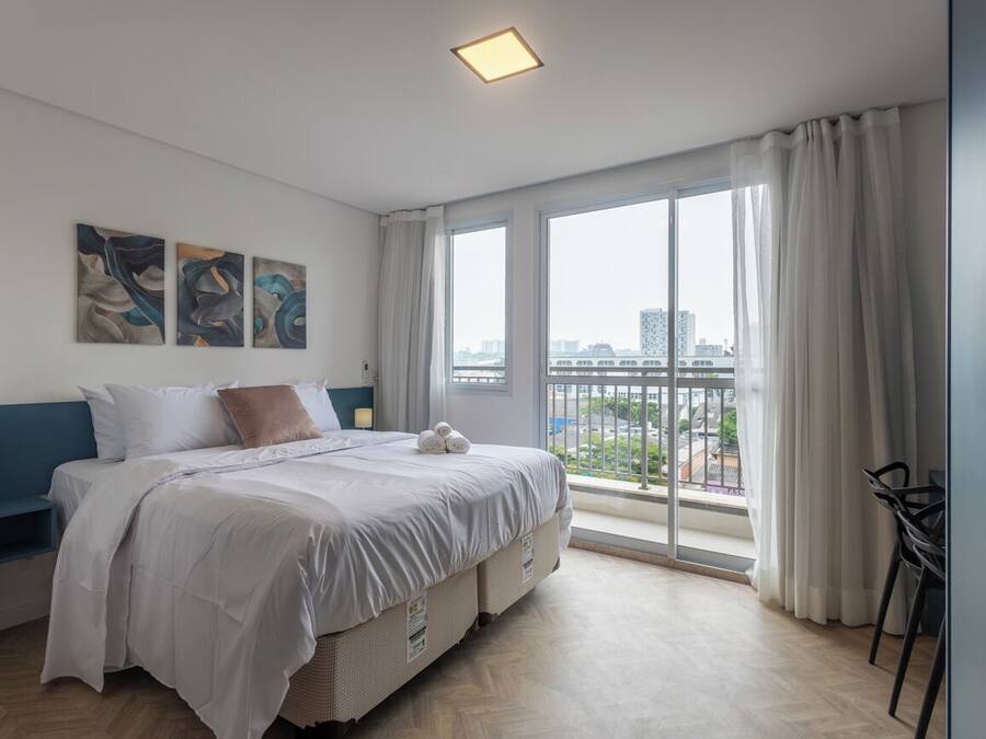 Quarto moderno com uma cama grande, vista para a cidade e uma varanda com uma cadeira.