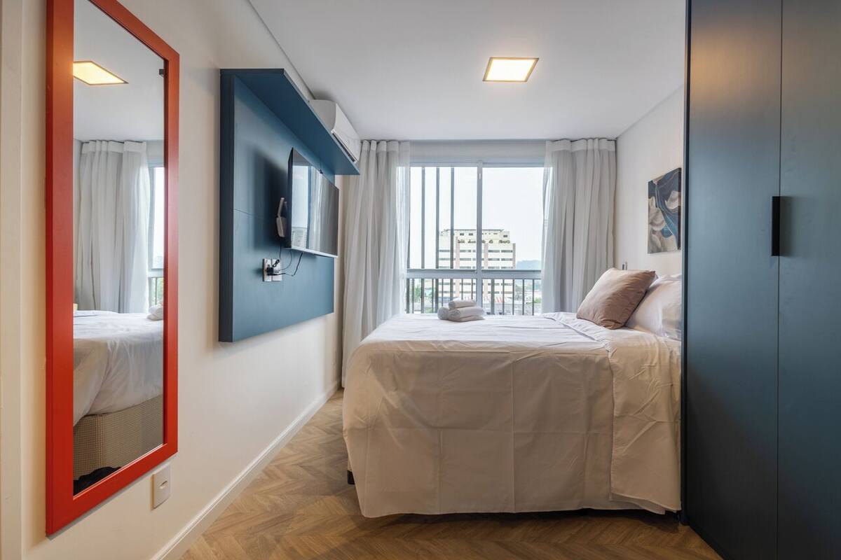 Une chambre d’hôtel avec un lit, un grand miroir, un téléviseur fixé au mur et une fenêtre avec des rideaux.