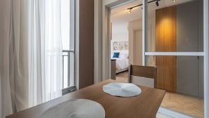 Une chambre d’hôtel moderne avec une table à manger en bois, une grande fenêtre avec des rideaux légers et vue sur la chambre.