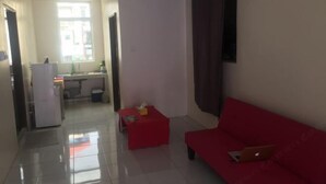 A room with a red sofa, a small red table, a white air conditioning unit, and a kitchen area in the background.