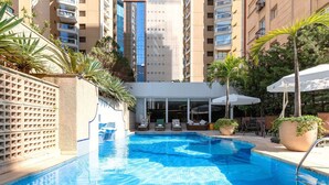 A swimming pool with lounge chairs and umbrellas, surrounded by tall buildings and greenery. - Get a Flat 1910 - Jardim Paulista (São Paulo)