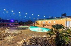A swimming pool with a surrounding deck, a building with a flat roof, and string lights overhead.