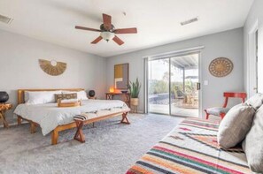 A bedroom with a bed, a ceiling fan, a large window, and a view of the outdoors. - Yucca Valley Oasis: Pool, Spa & Game Room Retreat (Yucca Valley)
