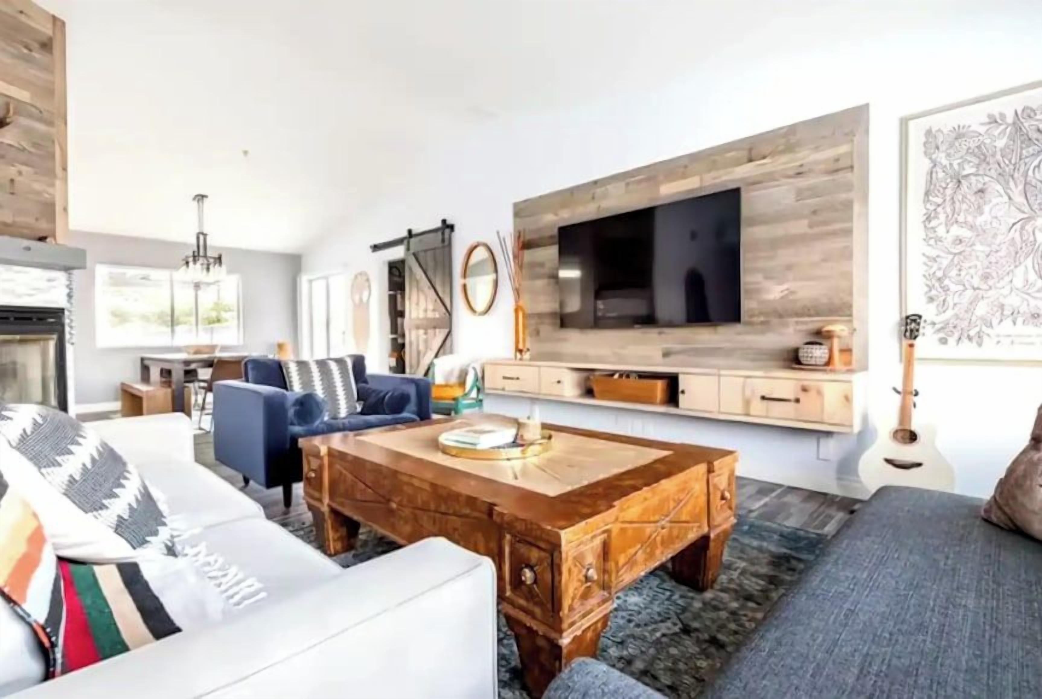 A modern living room with a fireplace, a large TV mounted on a wooden wall, a sofa, a coffee table, and a guitar.